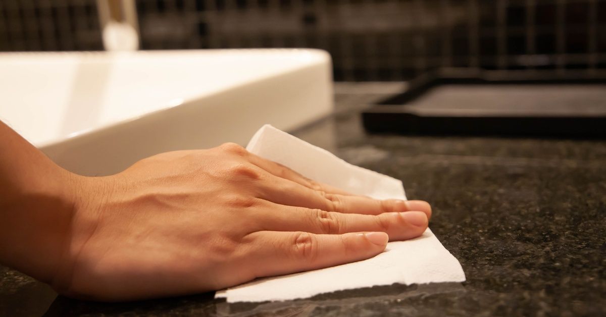 A close-up of a person's hand using a folded paper towel to wipe up liquid on a granite countertop next to a bathroom sink.
