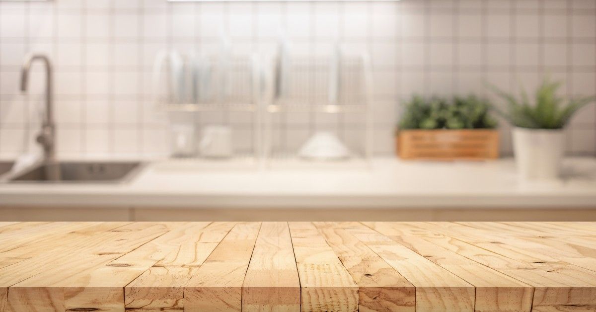 A wooden countertop in a light color. In the background is a blurry sink, salt and pepper shakers, and small plants.