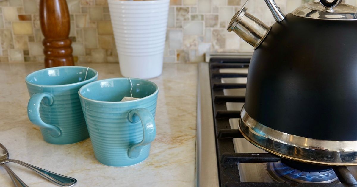Two blue mugs with tea bags in them. The mugs are on a quartzite countertop next to a stove with a teapot on it.