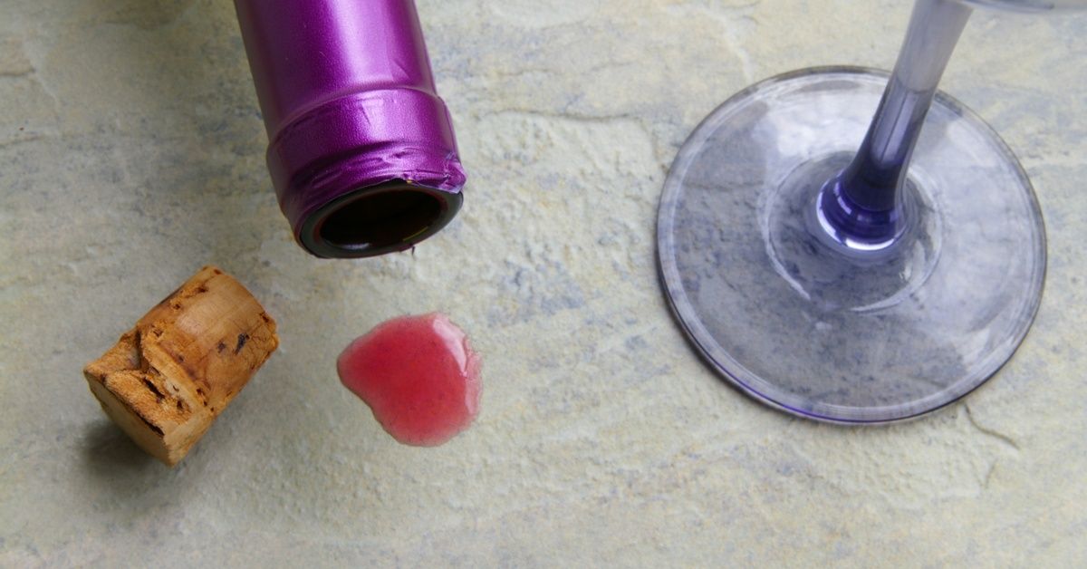 A purple wine bottle overturned next to a glass and a cork on a countertop. A red stain is visible.