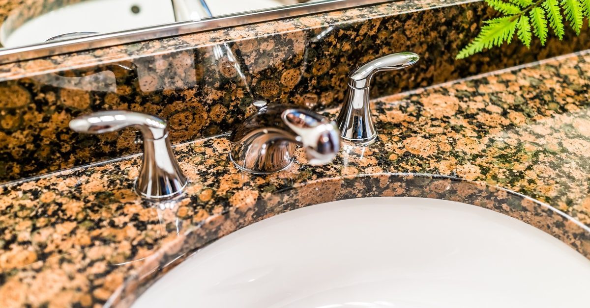 A bathroom countertop made of brown granite. The counter has a white sink and a silver faucet installed on its surface.