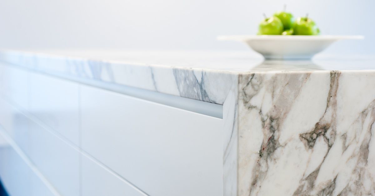 A white bowl full of green apples sitting on a stone countertop. The countertop has a waterfall style edge.