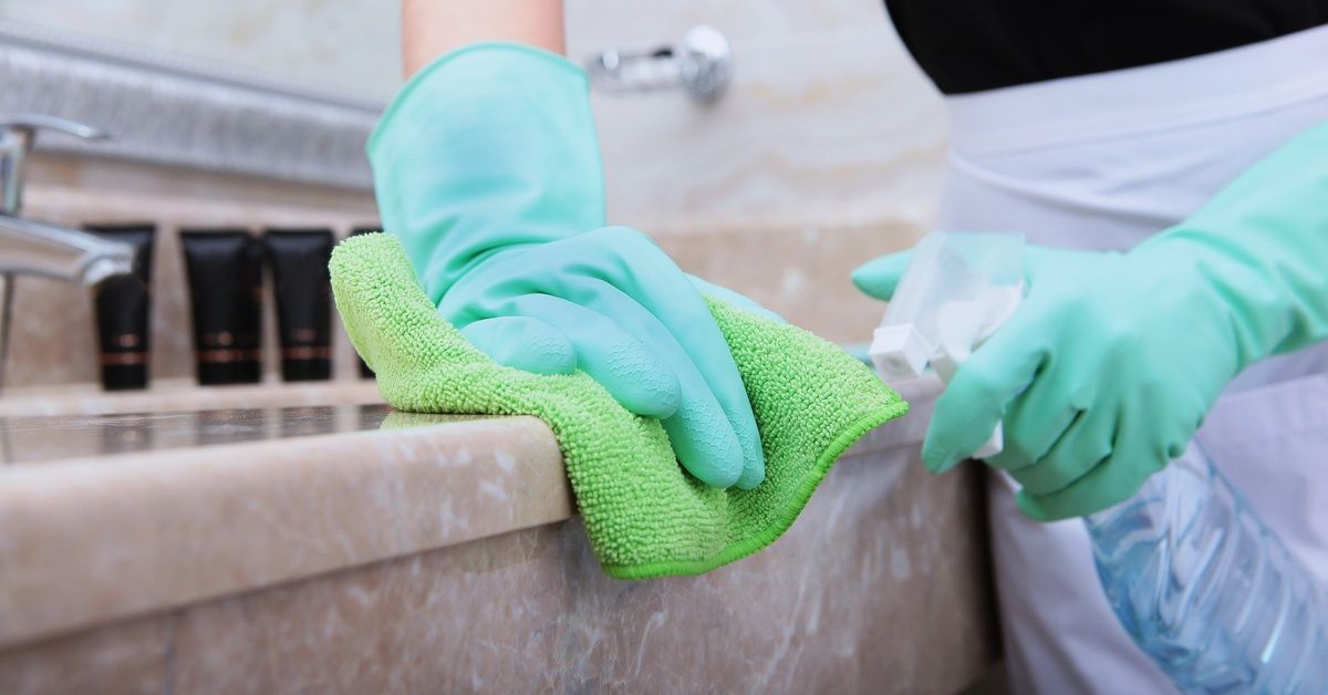 A person wearing bright green gloves is using a spray bottle and rag to clean the edge of a stone countertop.