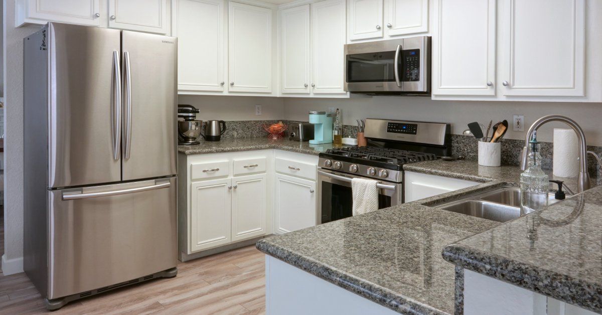  A brightly lit kitchen with white wooden cabinets and granite countertops. The floor is made of wooden planks.