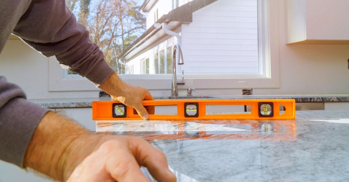 A person holding an orange level on a sleek stone countertop near a large window in a residential kitchen.