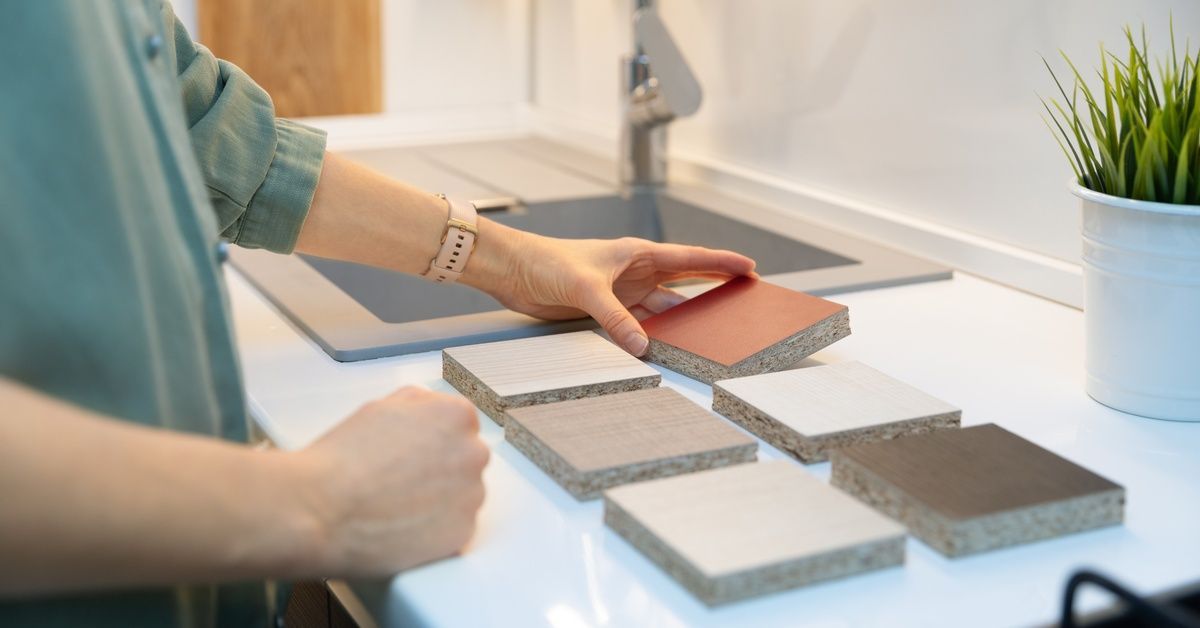 Six countertop material samples on a white kitchen counter. A person reaches for one of the samples.