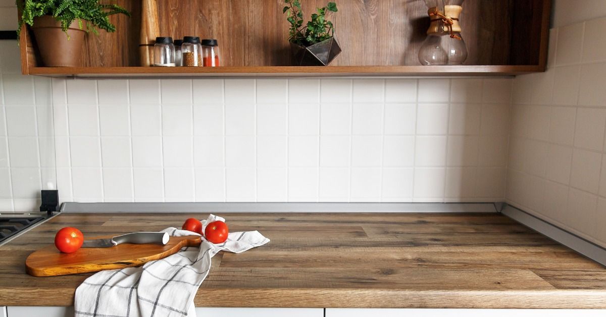 A wood countertop in a kitchen. Three tomatoes, a knife, a cutting board, and a white rag are on top of the counter.