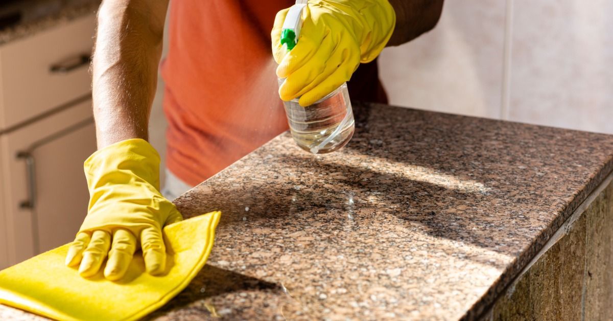 A person wearing yellow gloves using a yellow rag and spray bottle to clean a granite kitchen countertop.