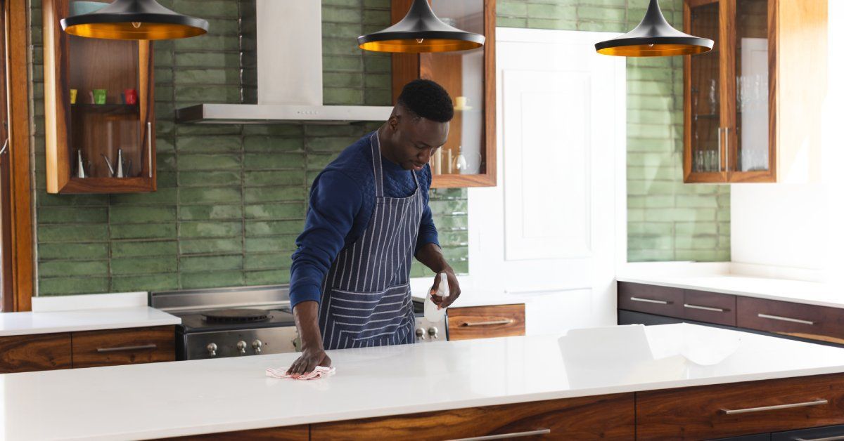 A Black male homeowner in his modern kitchen, wearing a blue apron as he wipes down his quartz countertops.