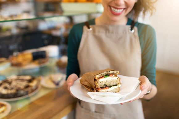 Smiling woman in apron holds a sandwich on a plate in a cafe, with a display case and baked goods visible.