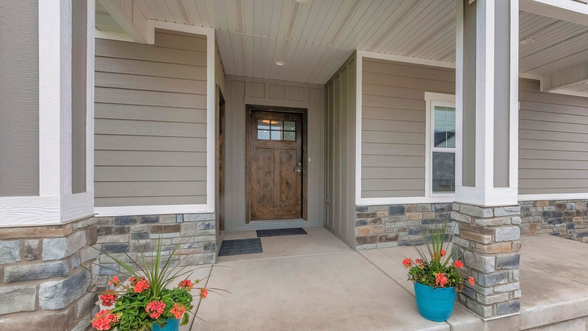 Front porch with stone columns, tan siding, a wooden front door, and two potted flowers.