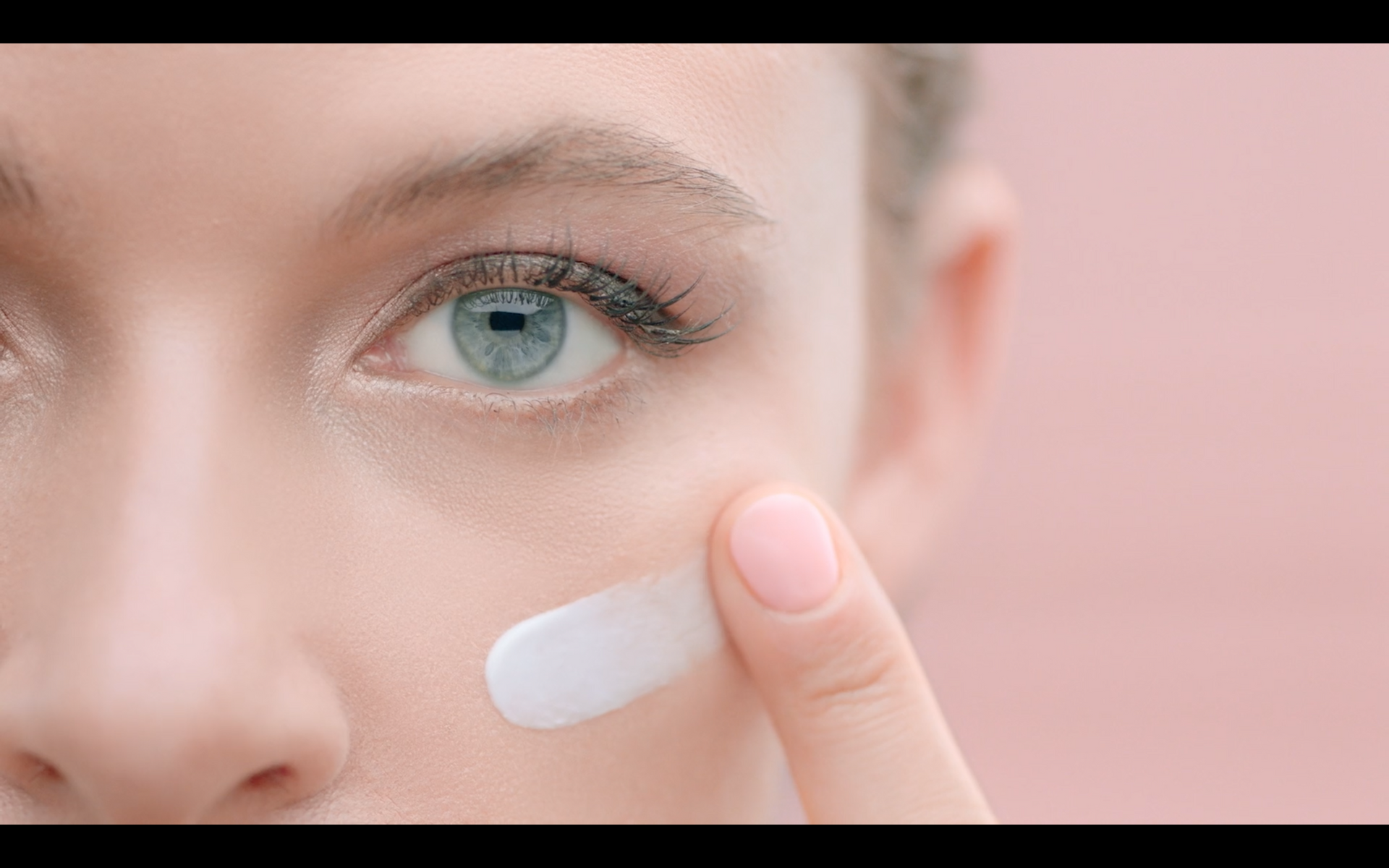 Close-up of a person applying cream near the eye; pale pink background.