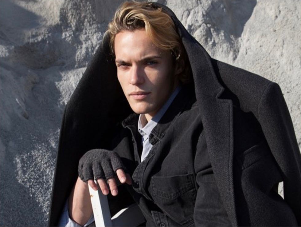 Blond-haired person in a black jacket and shirt, leaning against a light surface. Outdoors, neutral background.