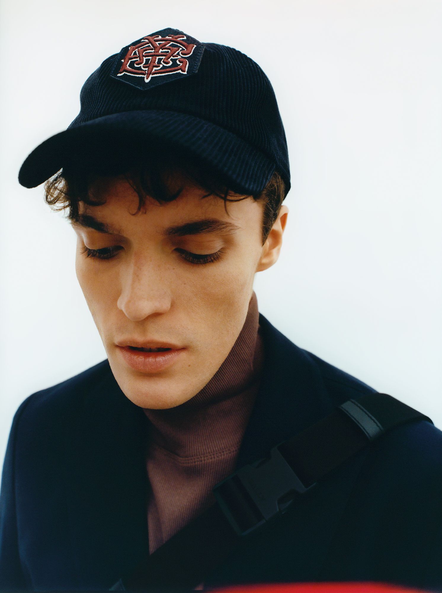 Man wearing a blue cap with a logo, looking down, wearing a brown turtleneck.