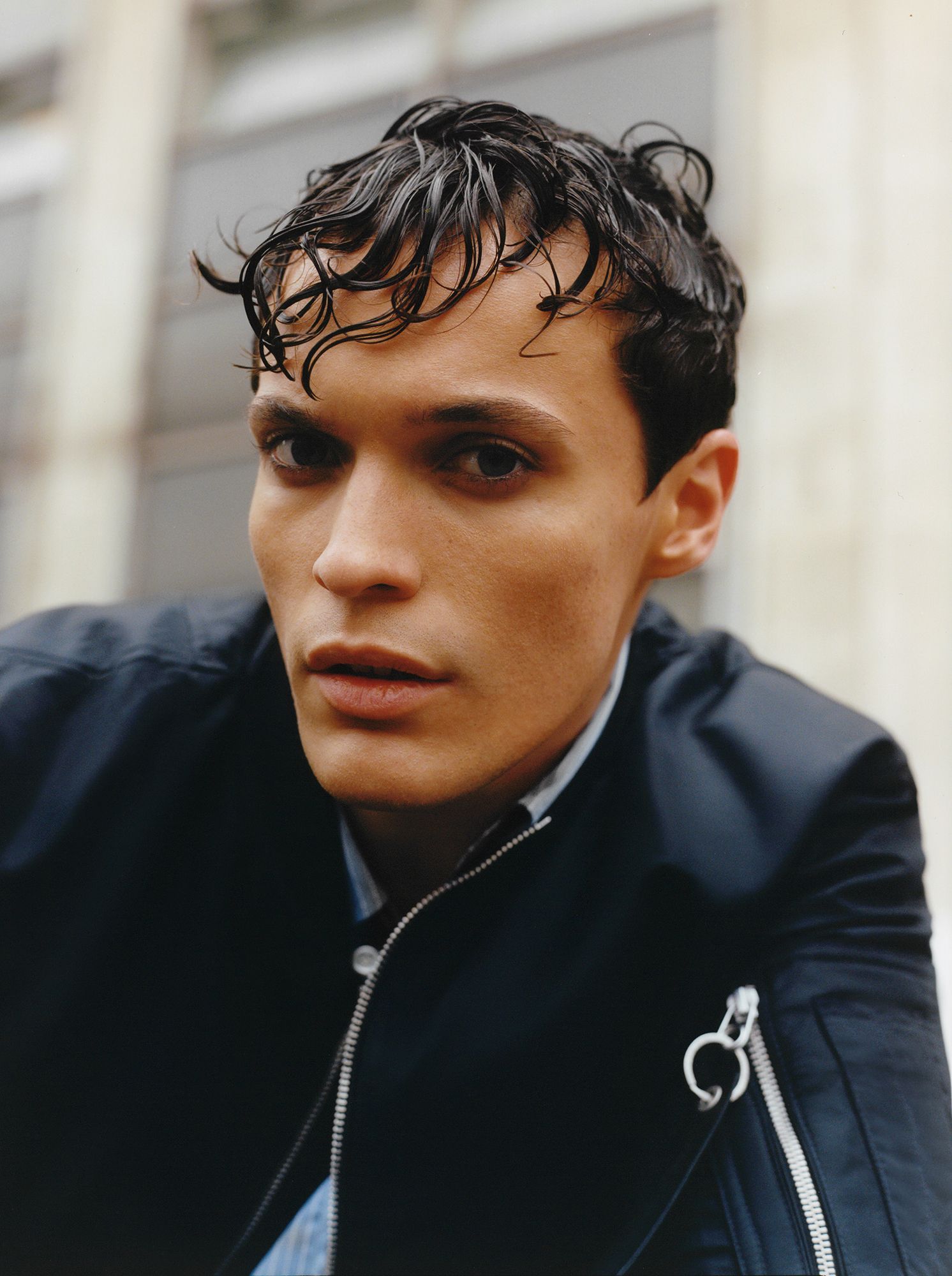 Man with wet, dark hair, wearing a black jacket, looking at the viewer. Outdoors with a building in background.