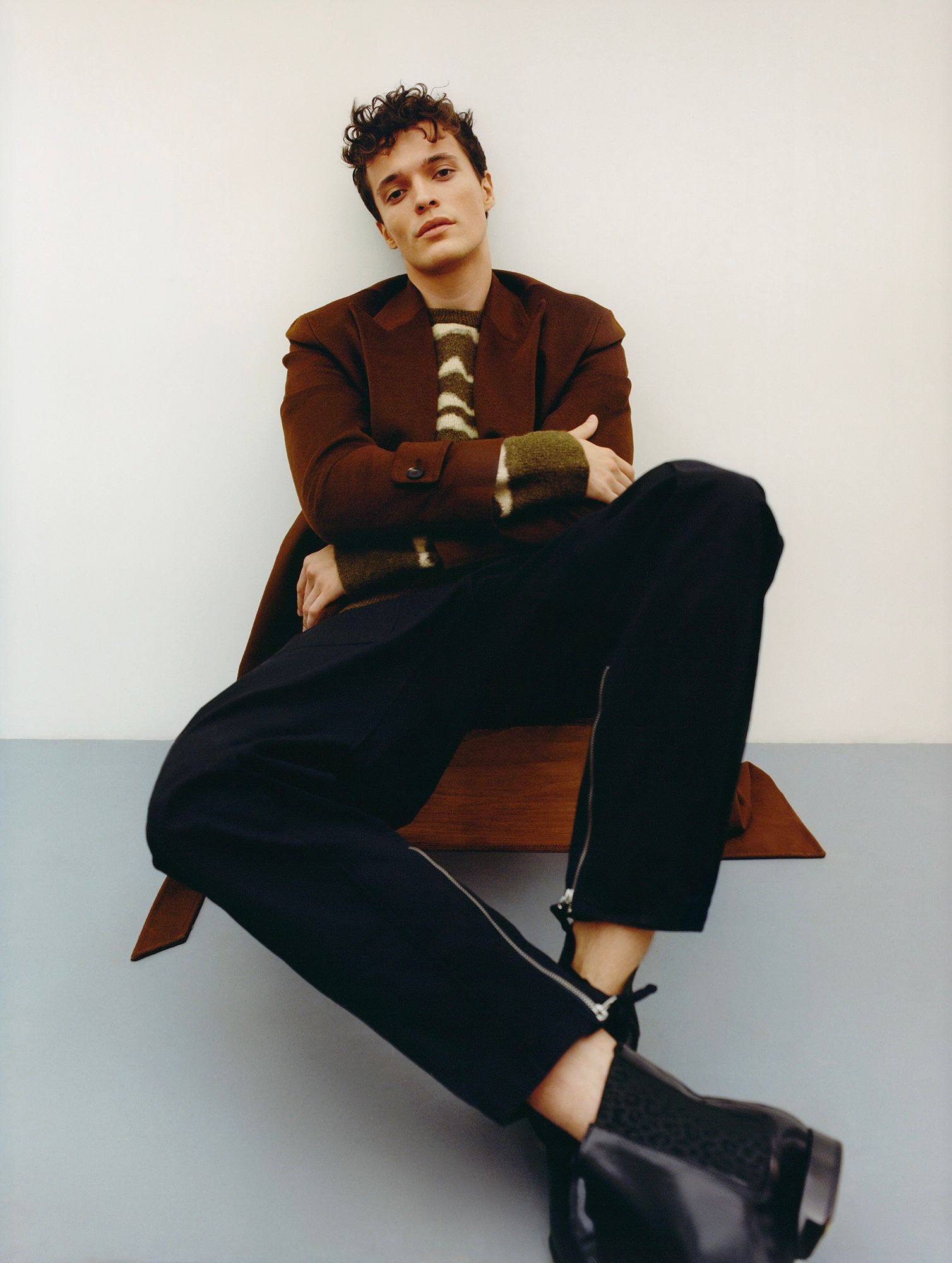 Man in brown blazer, striped sweater, and black pants sits on a wooden shelf, legs crossed.