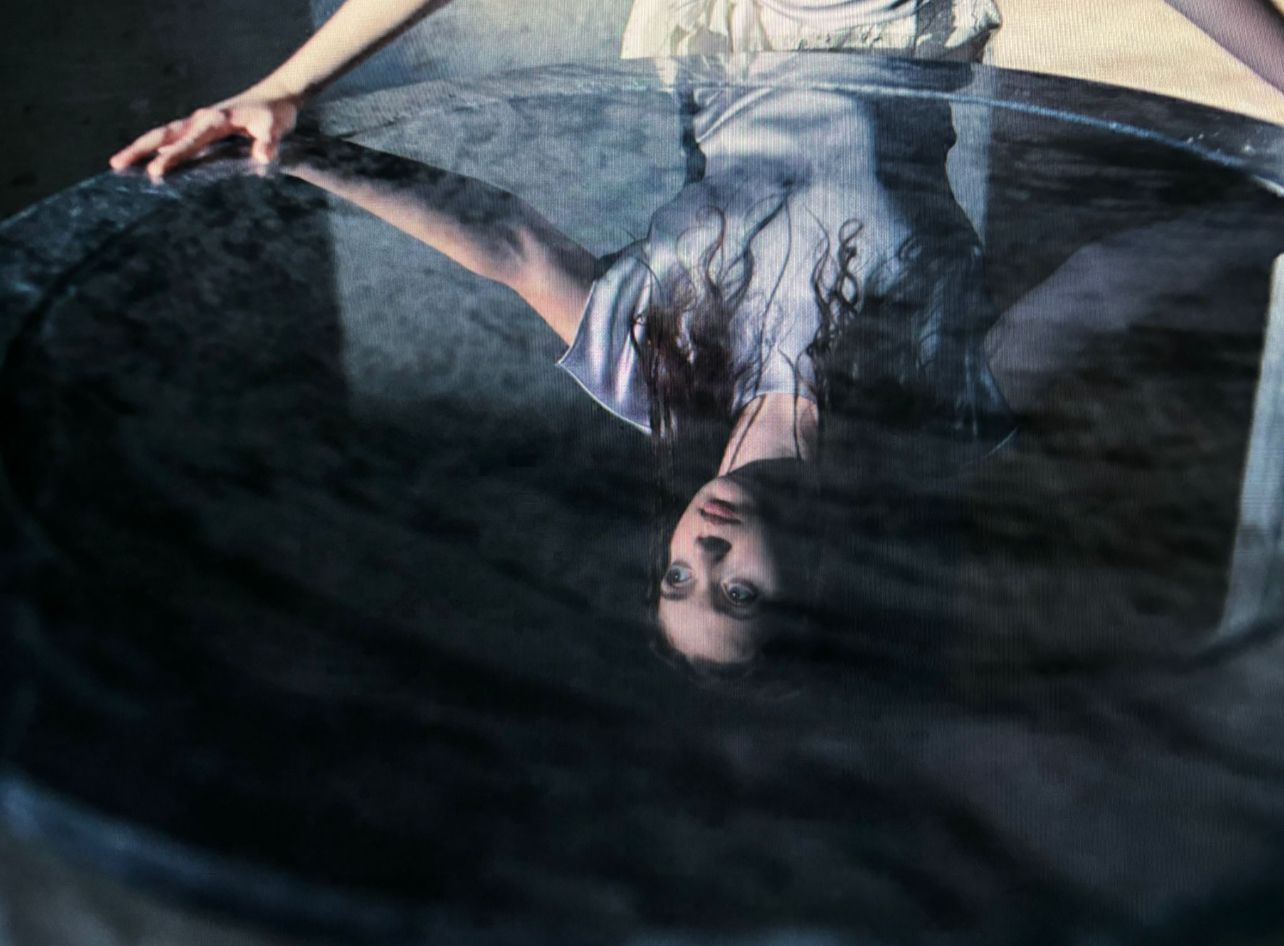 Woman reflected in dark water. Arms outstretched, she gazes up, wet hair, in a round, stone-like tub.