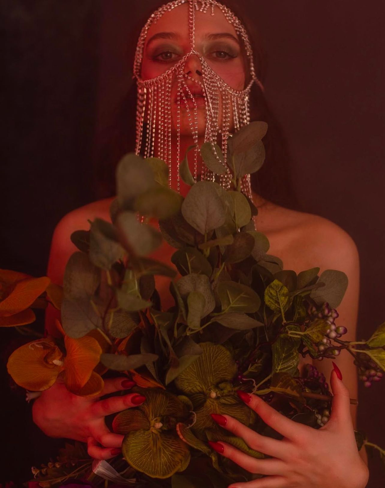 Woman wearing a jeweled face covering, holding a bouquet of green and red leaves, in dim lighting.