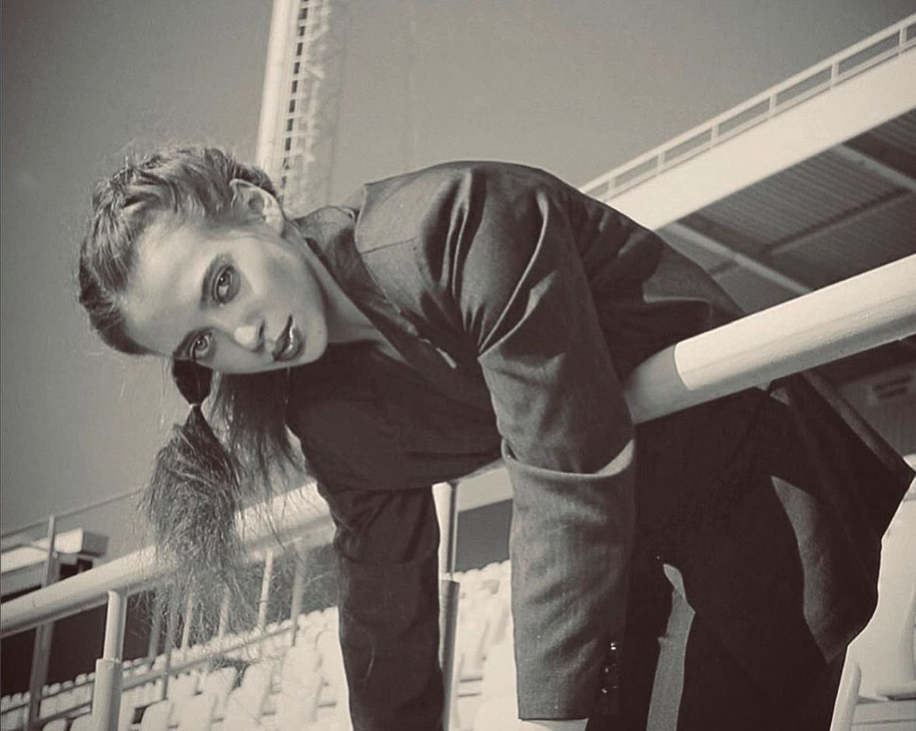 Woman in suit leans against stadium railing, looking toward the camera. Black and white.