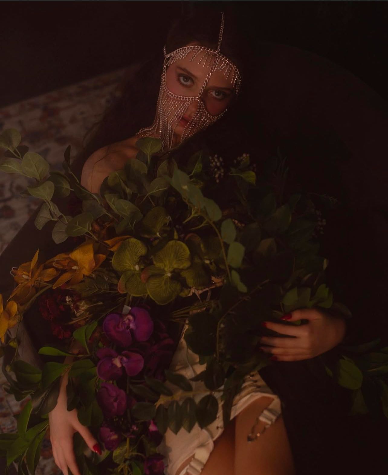 Woman in jeweled face covering holds bouquet of flowers; dim, moody lighting.