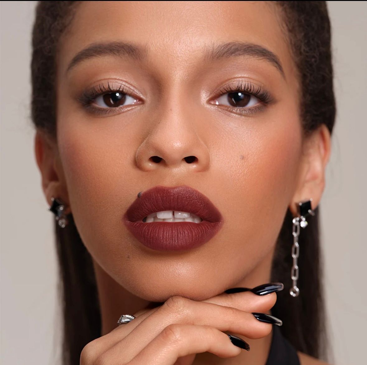Woman with dark lipstick and black nails, wearing earrings, holding chin.