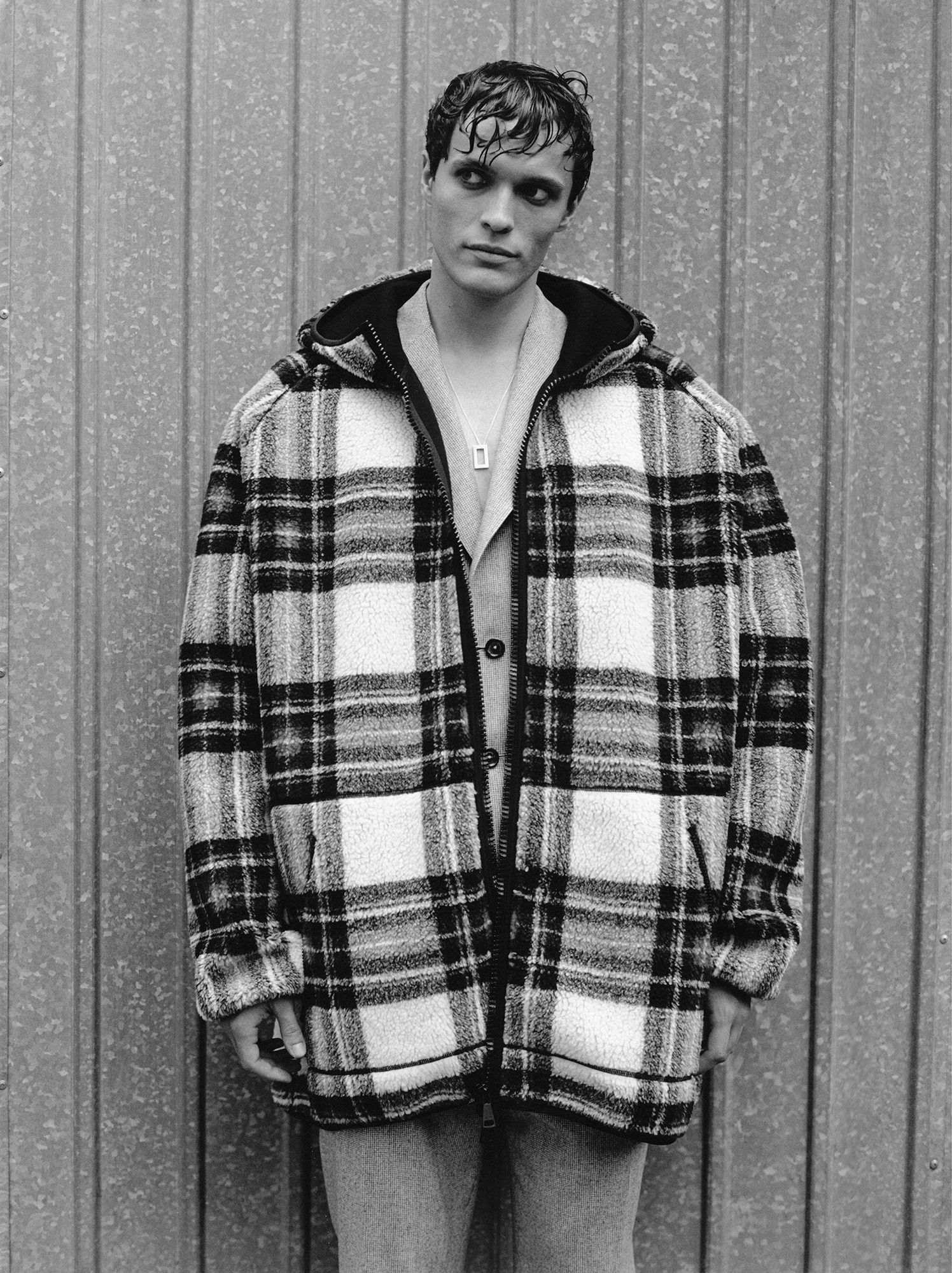 Man in plaid hooded jacket standing against a corrugated wall.