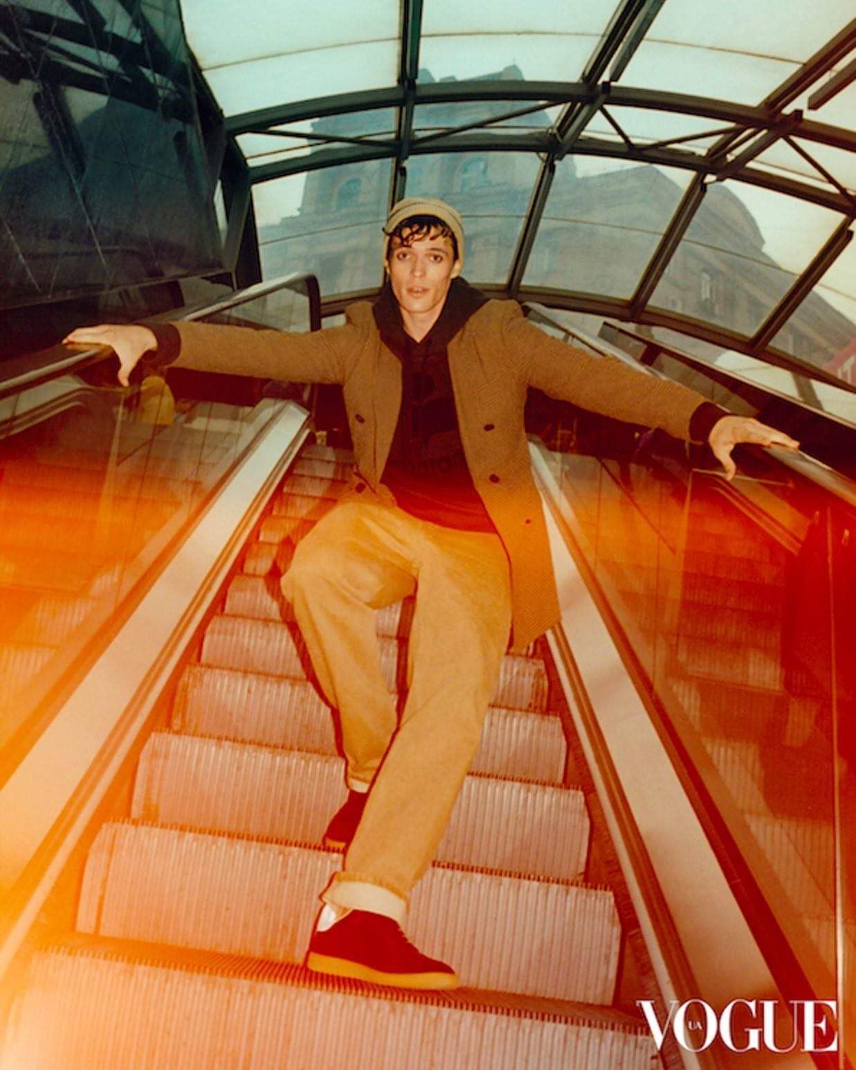 Man sitting on an escalator wearing a coat and cap, arms outstretched. VOGUE logo in the corner.