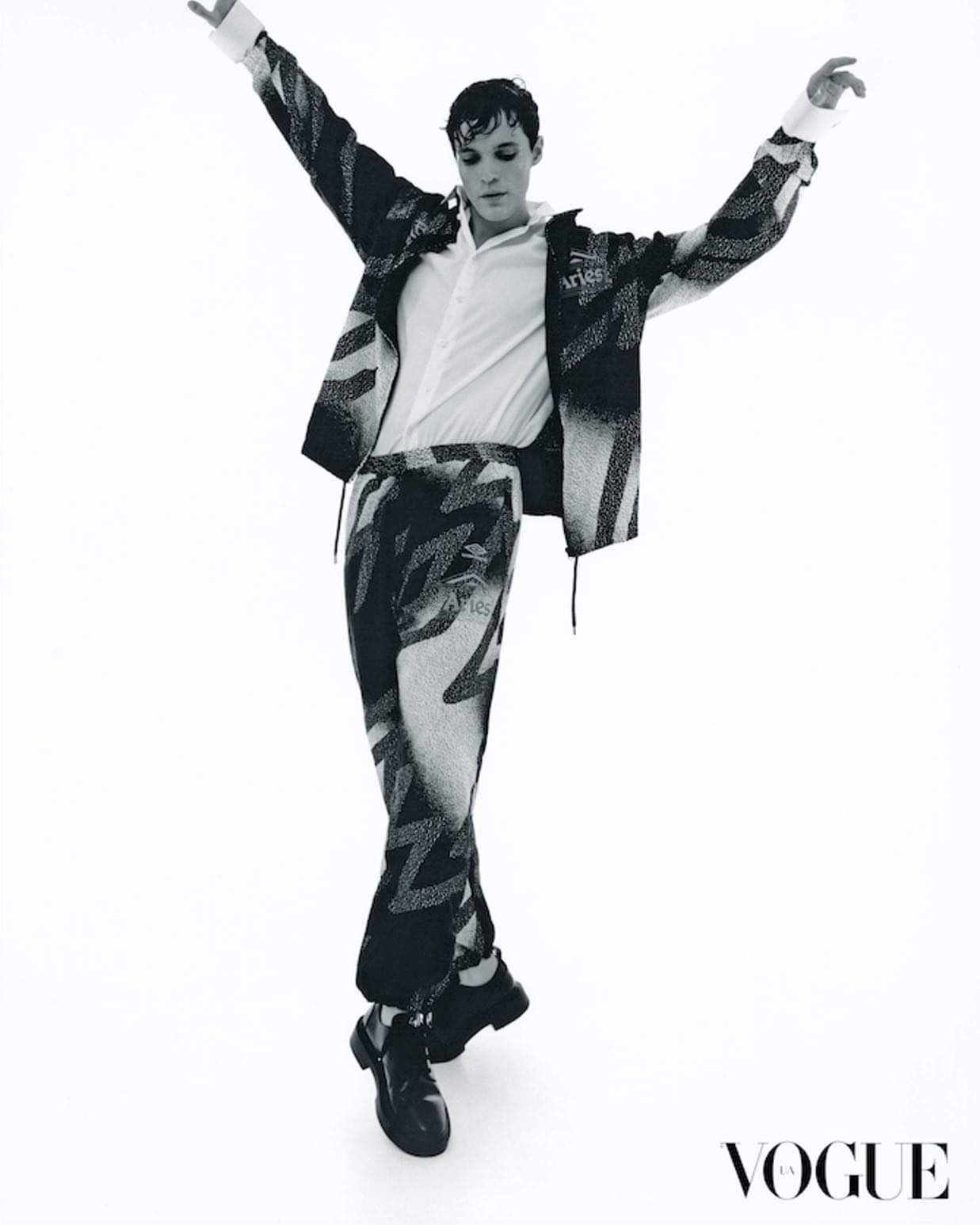 Person in patterned suit, arms outstretched, posing against a white background; Vogue logo in the corner.