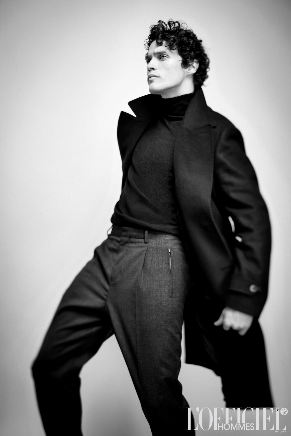 Man in black coat, turtleneck, and pants poses against white backdrop.