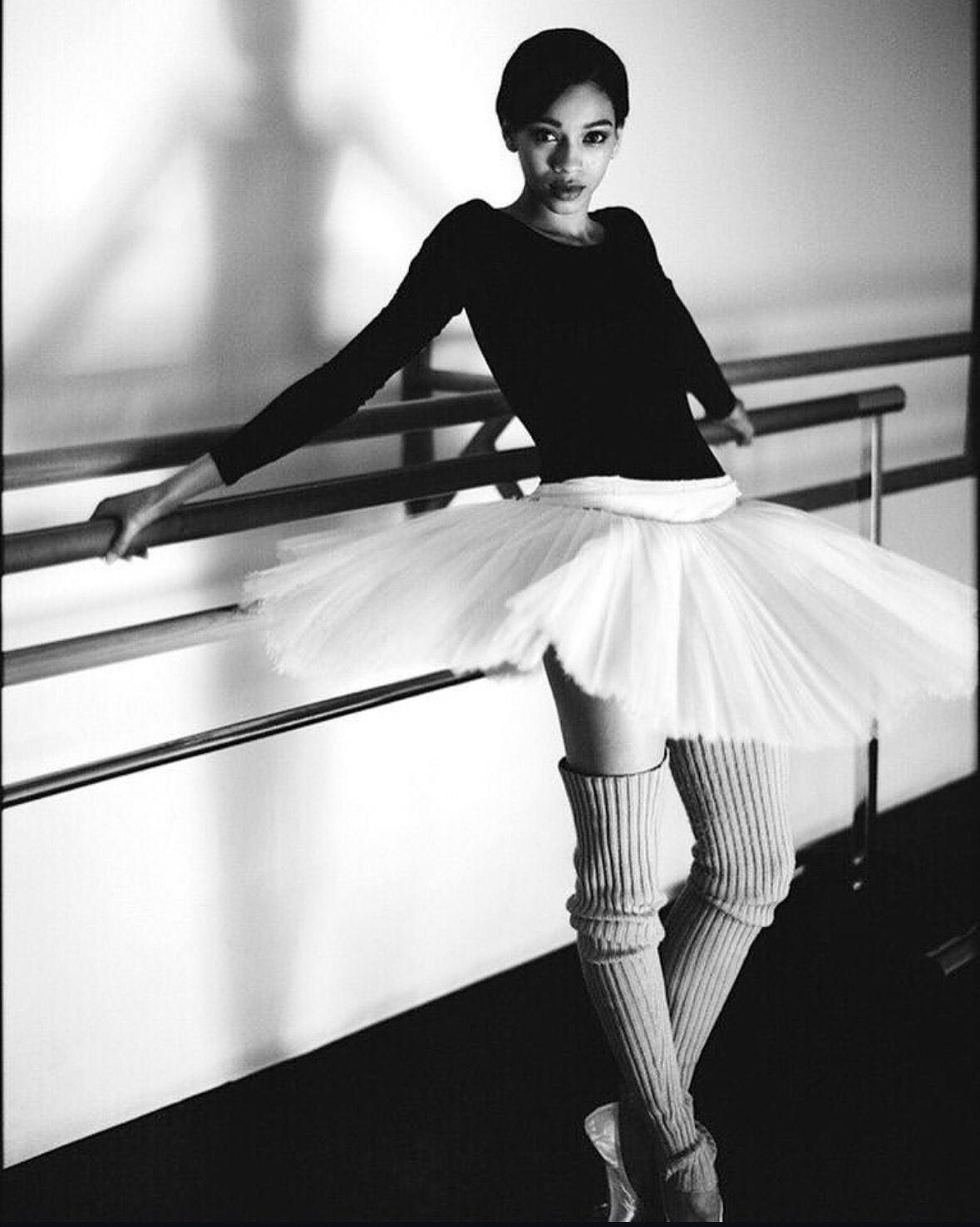 Ballerina in black leotard and white tutu, leaning on a ballet barre, wearing leg warmers.