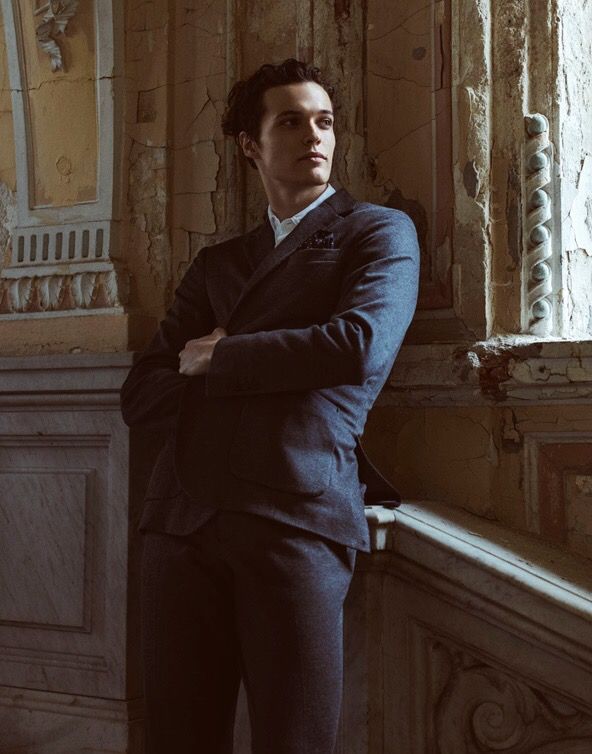Man in dark suit leans against decaying staircase in a crumbling building, arms crossed, looking away.
