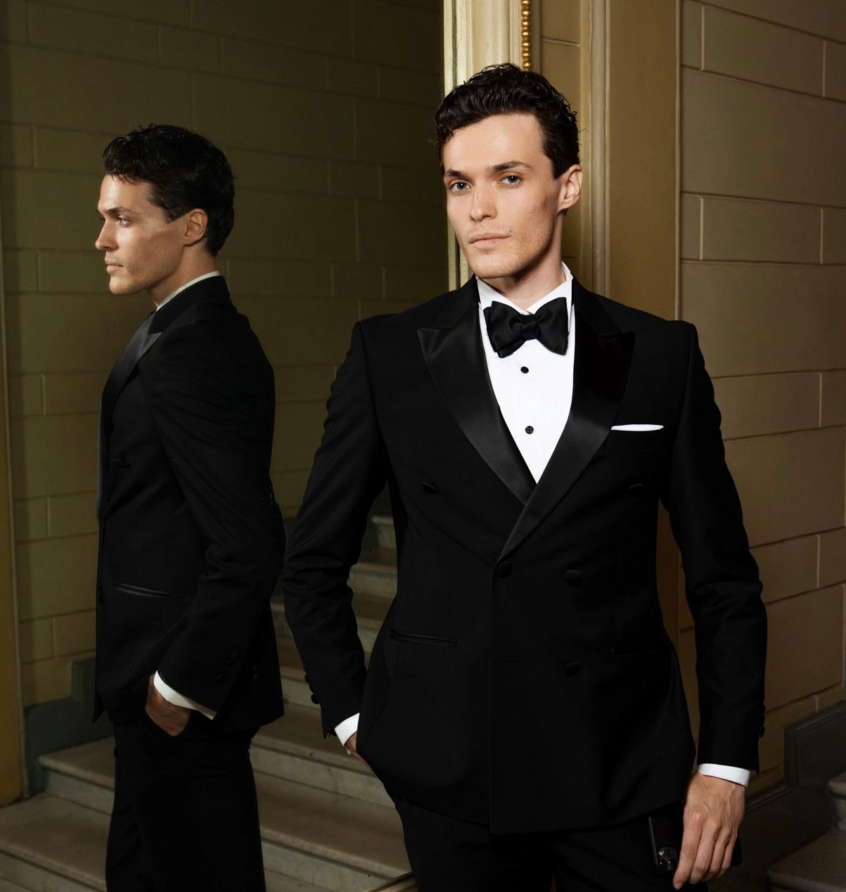 Man in a black tuxedo stands by a mirror, one hand in his pocket. Elegant interior with stairs.