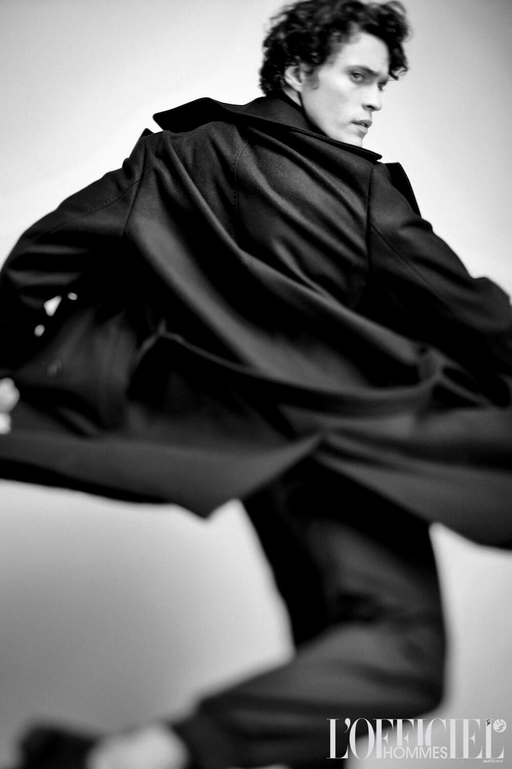 Man in black coat, mid-motion, blurred. Studio setting, L'Officiel logo.