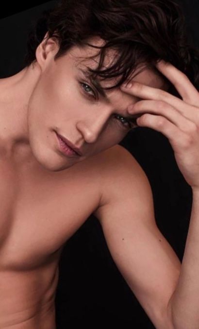 Man with dark wavy hair, leaning forward. Pale skin, focused expression, hand on forehead. Black background.