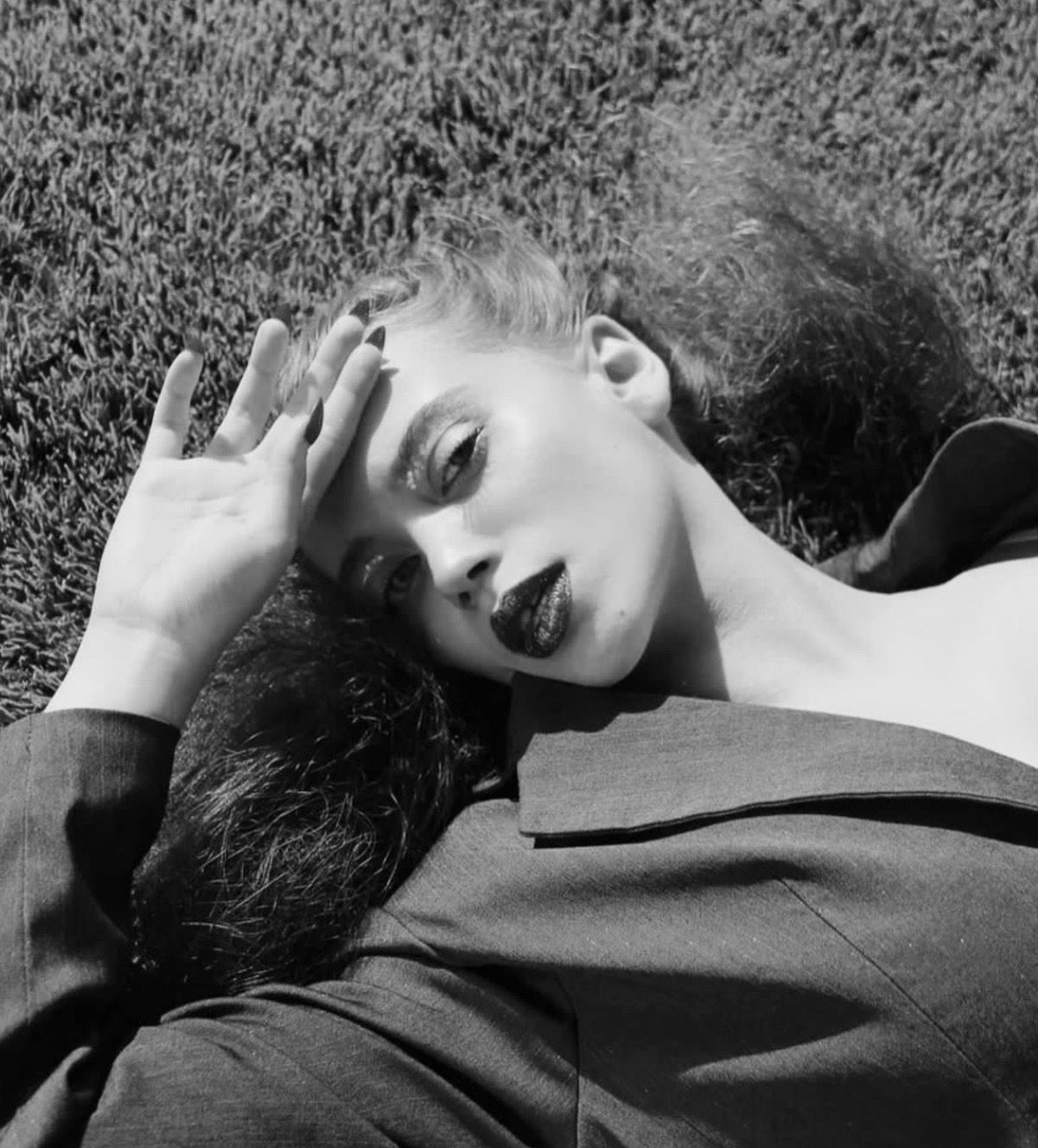 Woman reclines on grass, hand to forehead, wearing dark lipstick and blazer.