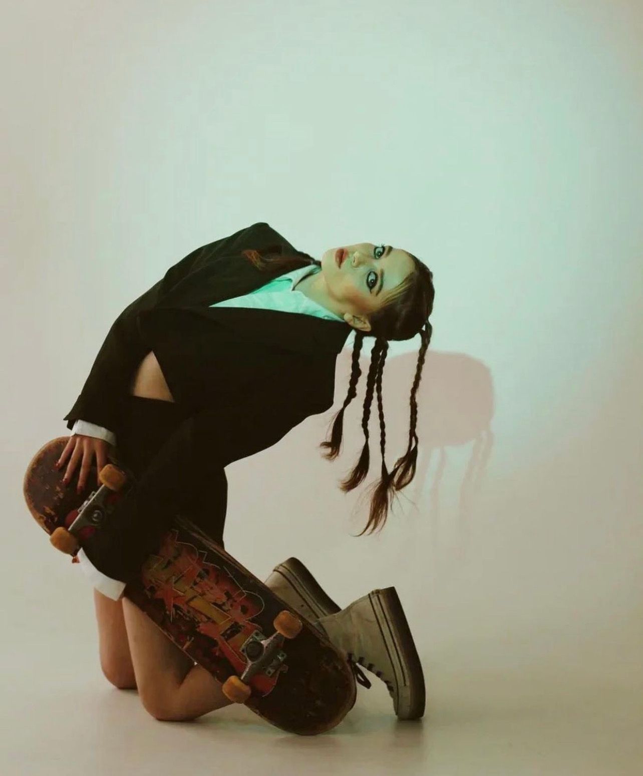 Person in suit arched backwards, kneeling with skateboards in hands, neutral background.