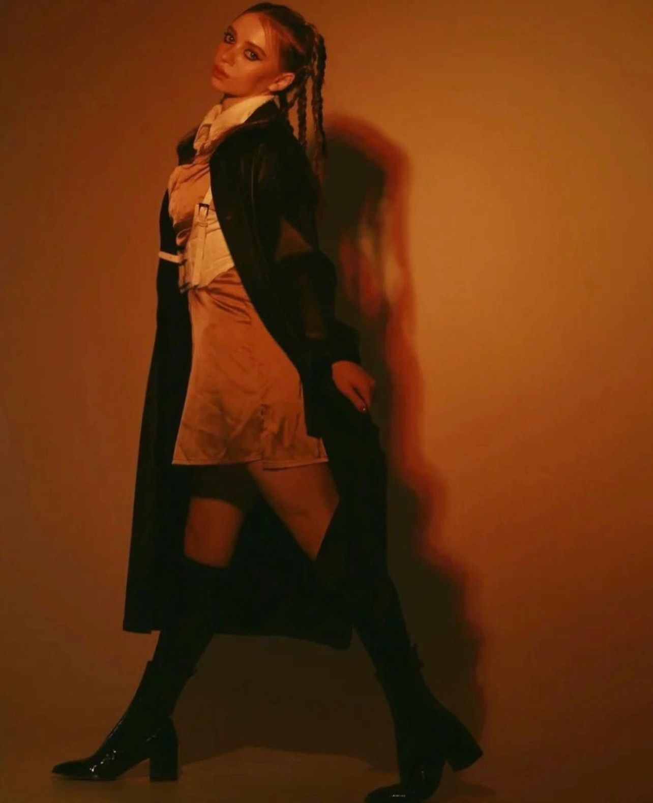 Woman in black coat and boots, walking, looking at camera. Golden light on peach background.