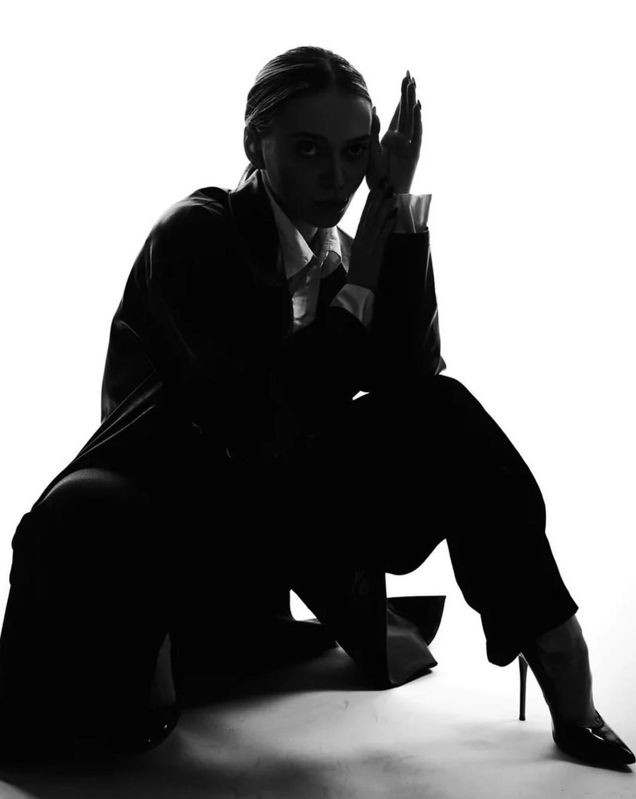 Woman in suit kneels, hands near face, looking toward the viewer in a black and white studio setting.