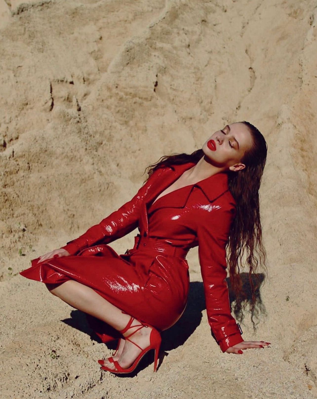 Woman in red patent leather outfit poses in front of a tan rock formation, with wet hair and red lipstick.