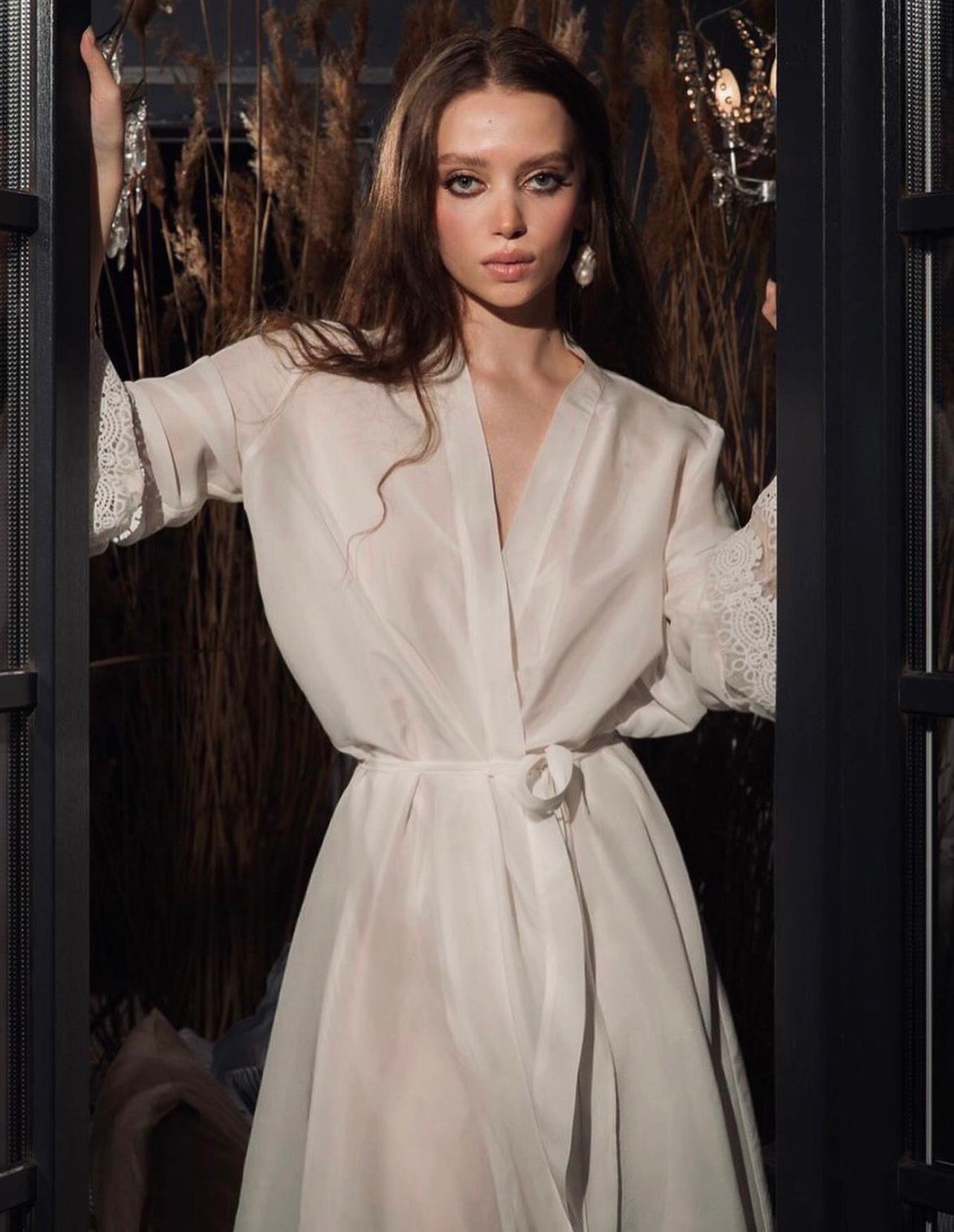 Woman in white robe, leaning in a doorway. She has long hair and wears dangling earrings. Dark background.