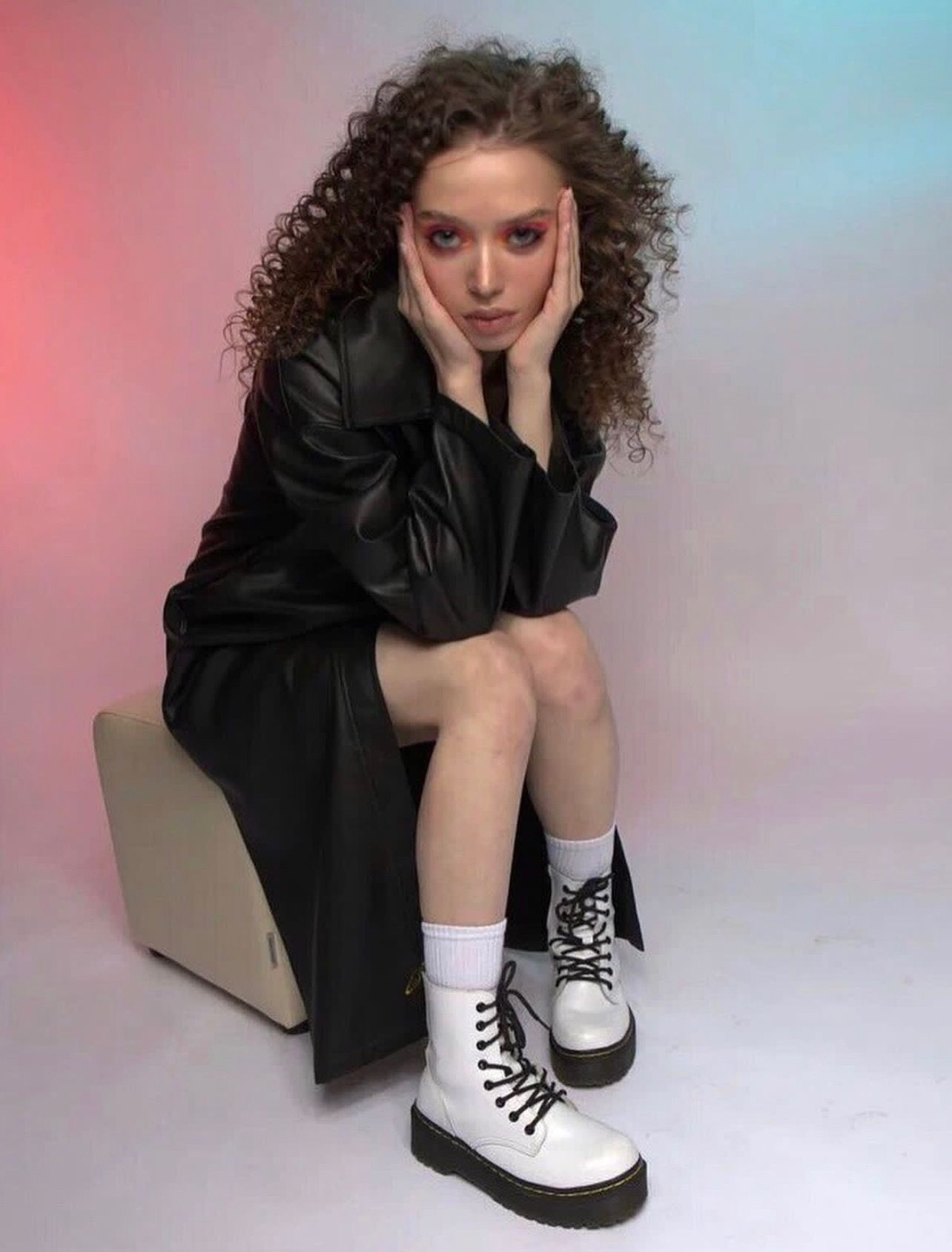 Woman with curly hair in black coat and white boots, resting chin on hands.