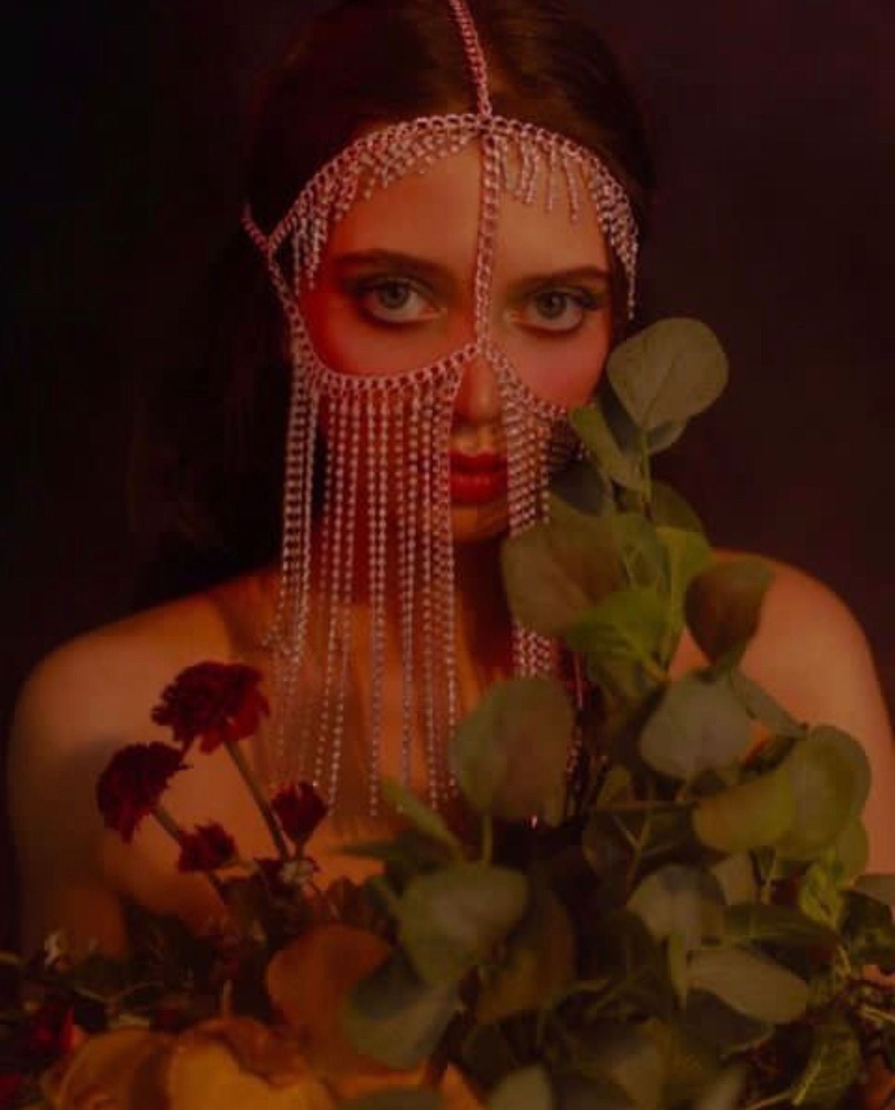 Woman wearing a chain mask and holding flowers against a dark red backdrop.