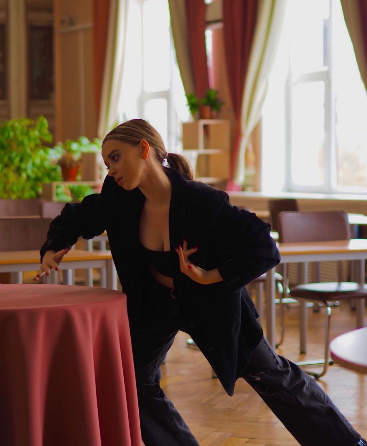 Woman in black suit dancing near a table in a room with windows and curtains.