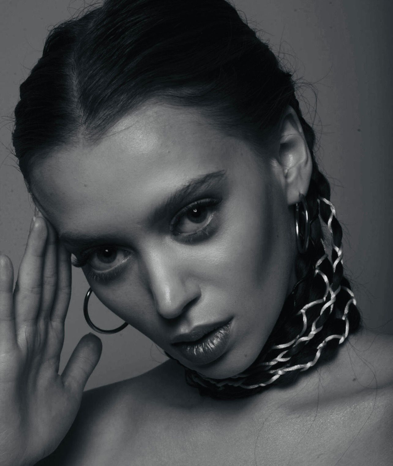 Woman with dark hair in a braid, wearing hoop earrings and a large neckpiece, gazing towards the camera.