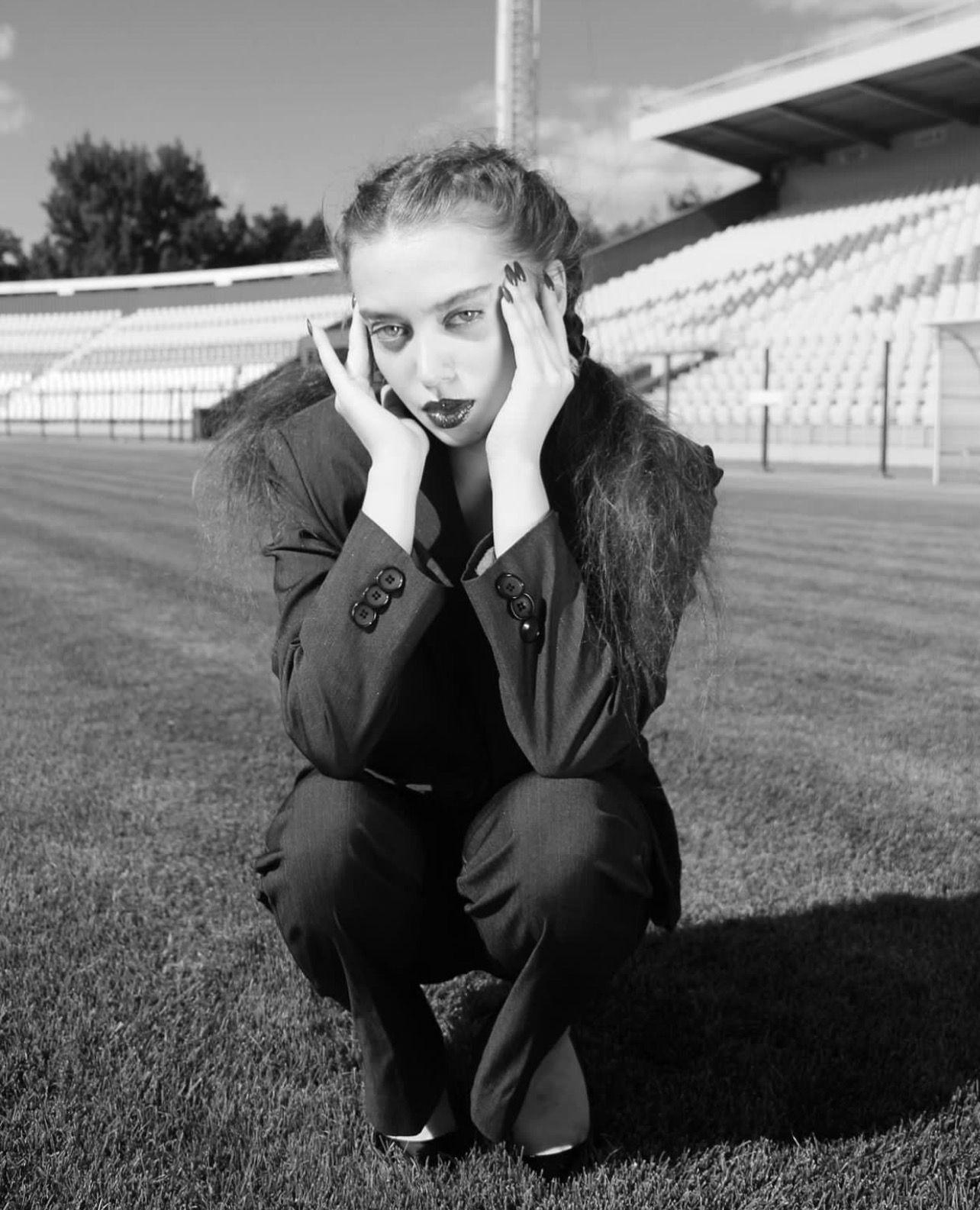 Person in dark suit squats on a soccer field, hands on their face, looking at the camera.