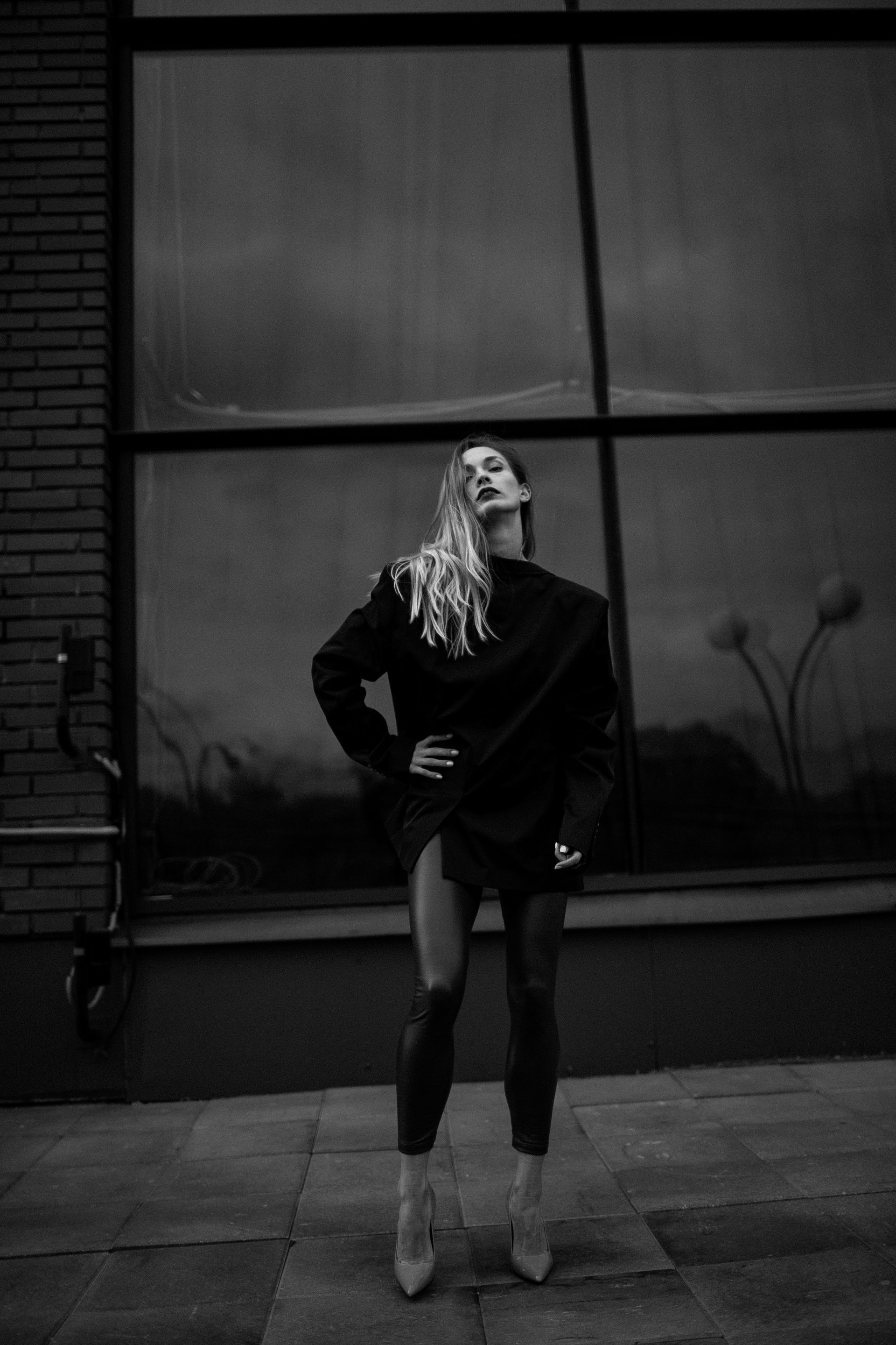 Woman in a black blazer and leggings, posed in front of a window. One hand on hip. Black and white.