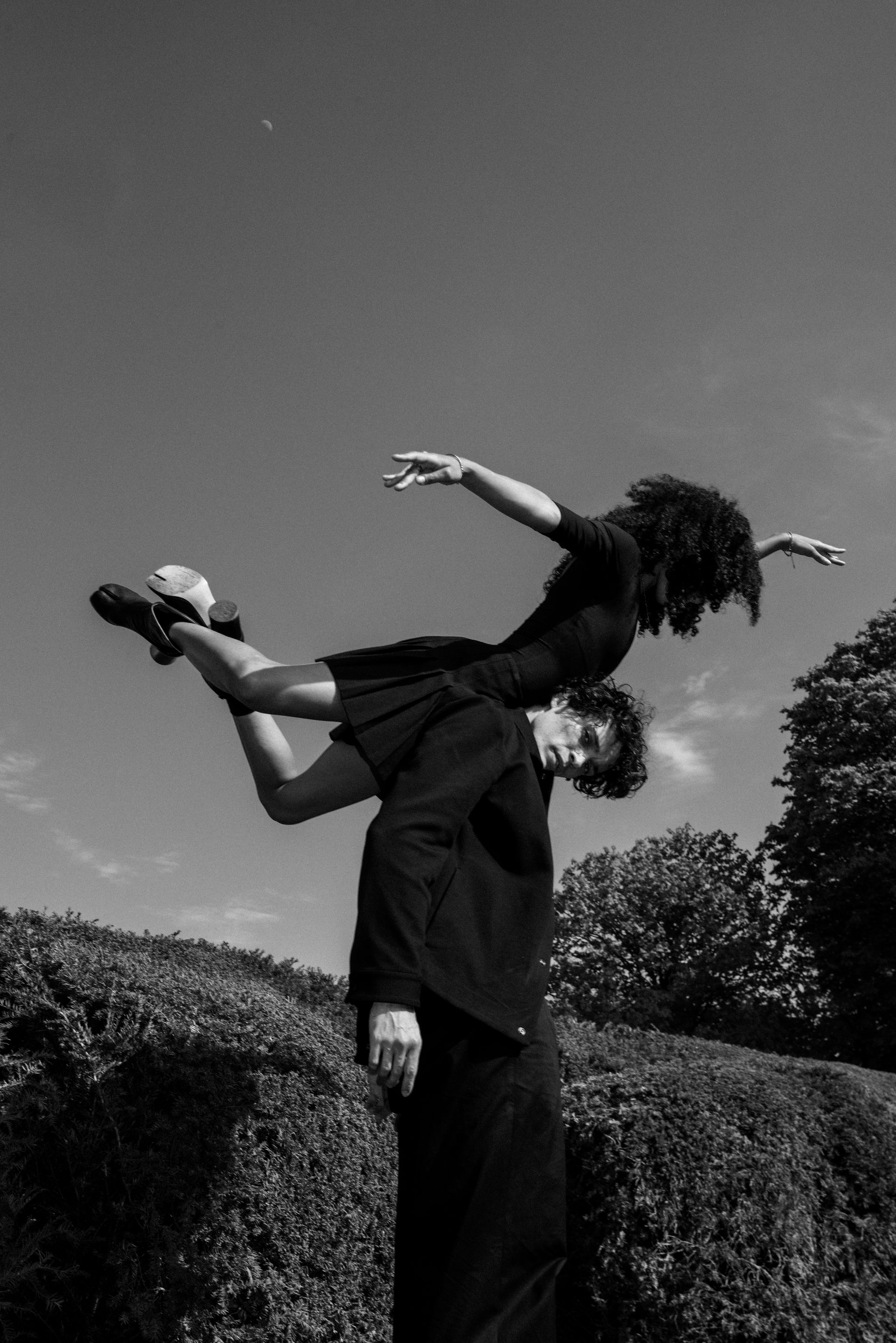 Man supporting a person as they balance overhead. Both are in dark clothing, set against a bright sky and manicured bushes.