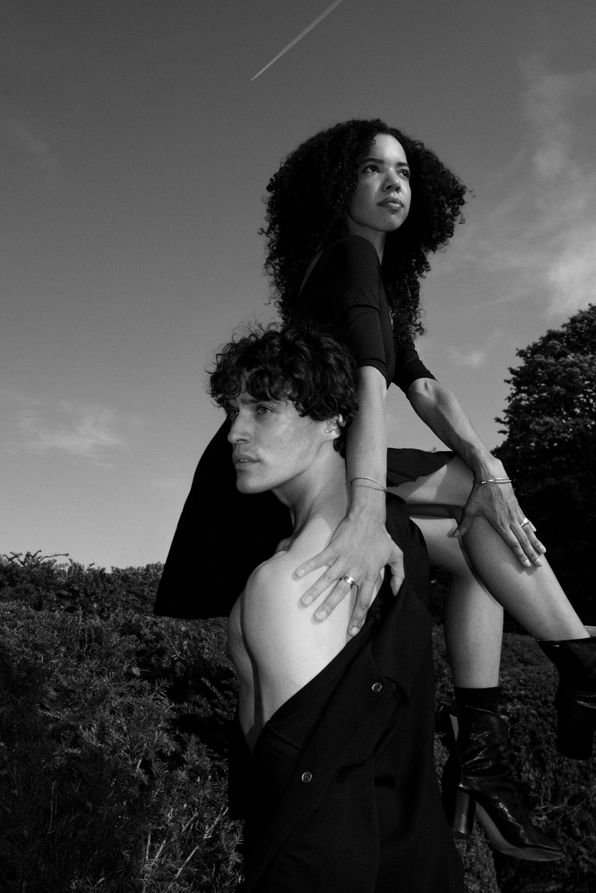 A person carries another on their shoulders, posing against a sky backdrop. Both wear black clothing.