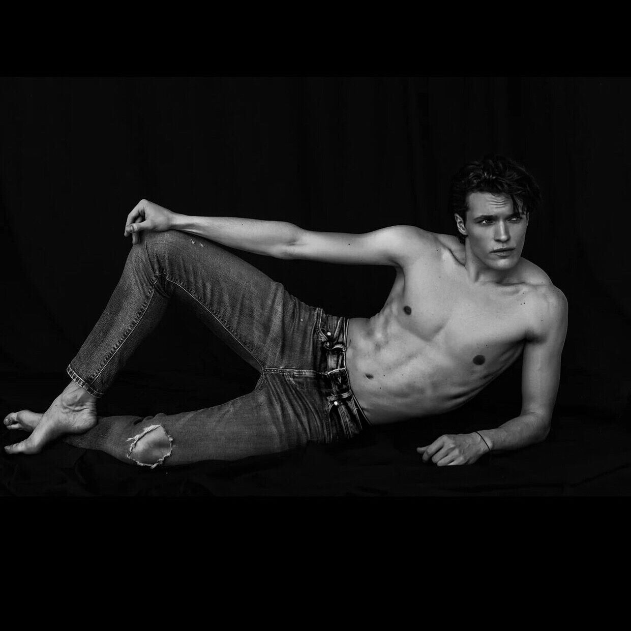 Man lying on his side in ripped jeans, shirtless. Black and white photo.