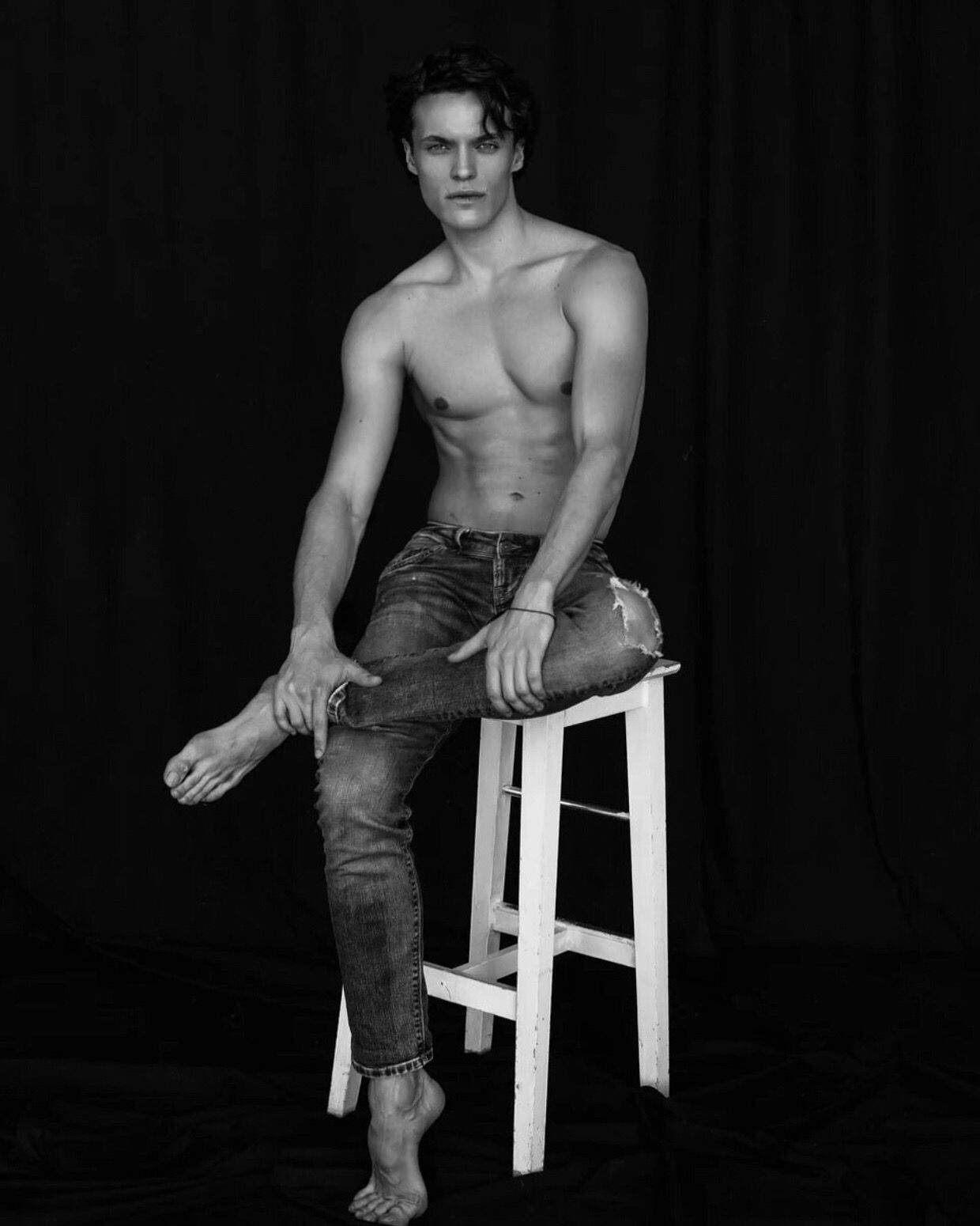 Man seated on stool, shirtless, in ripped jeans, barefoot, hand on foot, black background.